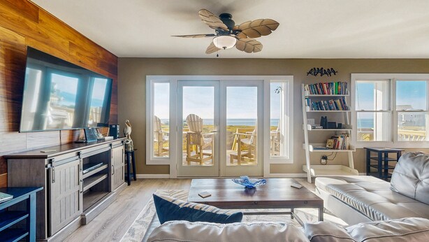 A spacious living room with a large window overlooking a balcony, a ceiling fan, and a bookshelf filled with books.