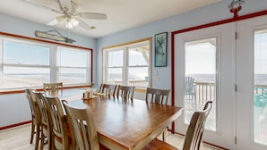 A dining room with a large wooden table and chairs, a ceiling fan, and large windows with a view of the beach. - Summer Sands: Waterfront, Dog Friendly, Balcony (Ocean Isle Beach)