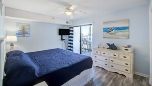 A bedroom with a bed, a dresser, a ceiling fan, a painting of a boat, and a view of the outdoors. - Oceanfront condo with elevator, private beach, ocean view, community pool, grill (North Topsail Beach)