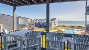 A covered outdoor area with white Adirondack chairs and a table, overlooking a pool and beach area. - Oceanfront condo with elevator, private beach, ocean view, community pool, grill (North Topsail Beach)