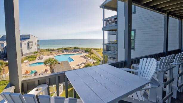 A balcony with a view of a pool and beach, white chairs, and a table. - Oceanfront condo with elevator, private beach, ocean view, community pool, grill (North Topsail Beach)