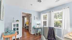 Quarto com piso de madeira, uma poltrona azul, uma pequena mesa com abajur e uma janela com cortinas brancas.