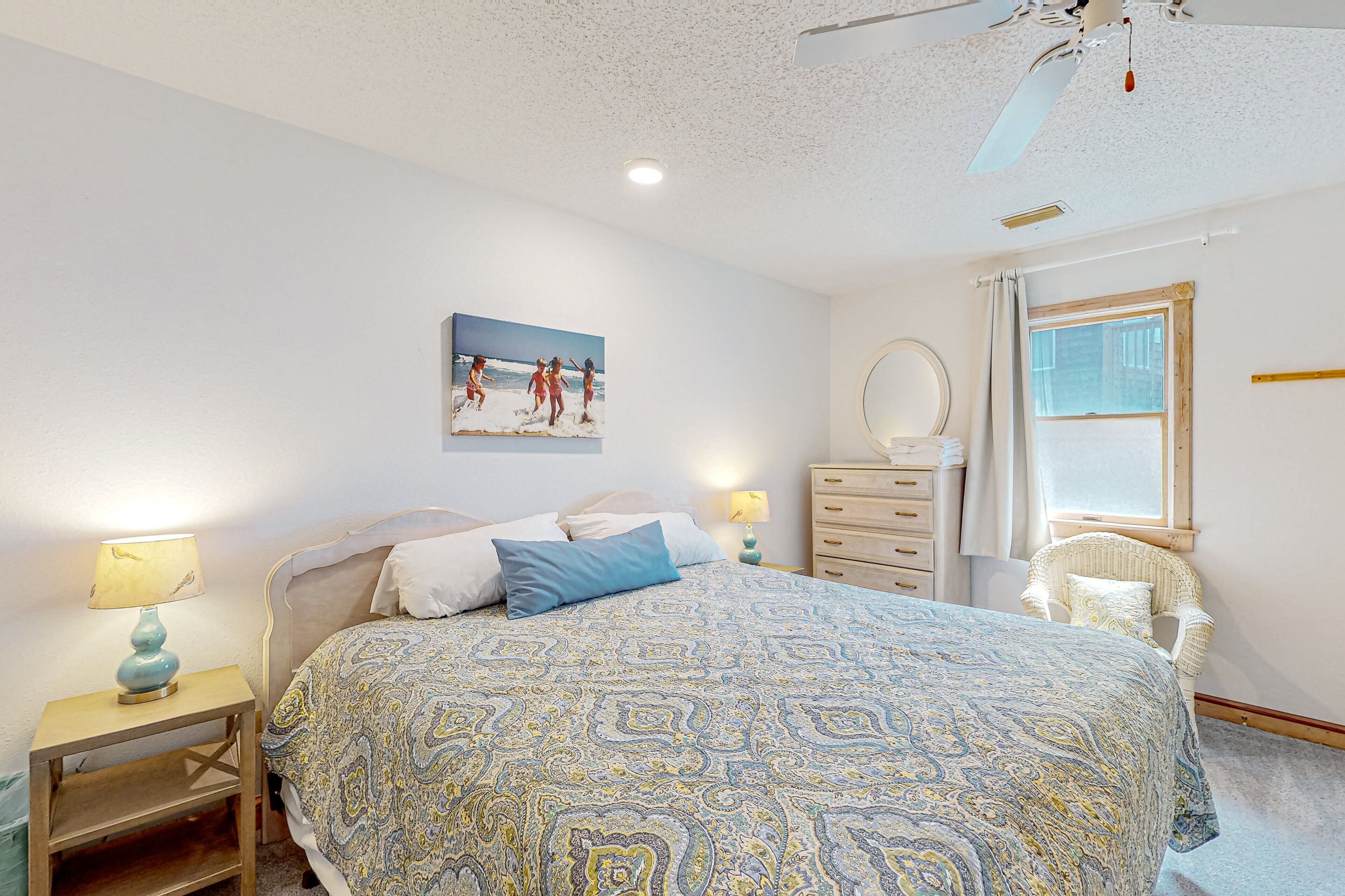 Une chambre à coucher comprenant un lit, une table de chevet, une commode, une chaise, une fenêtre et un ventilateur de plafond.