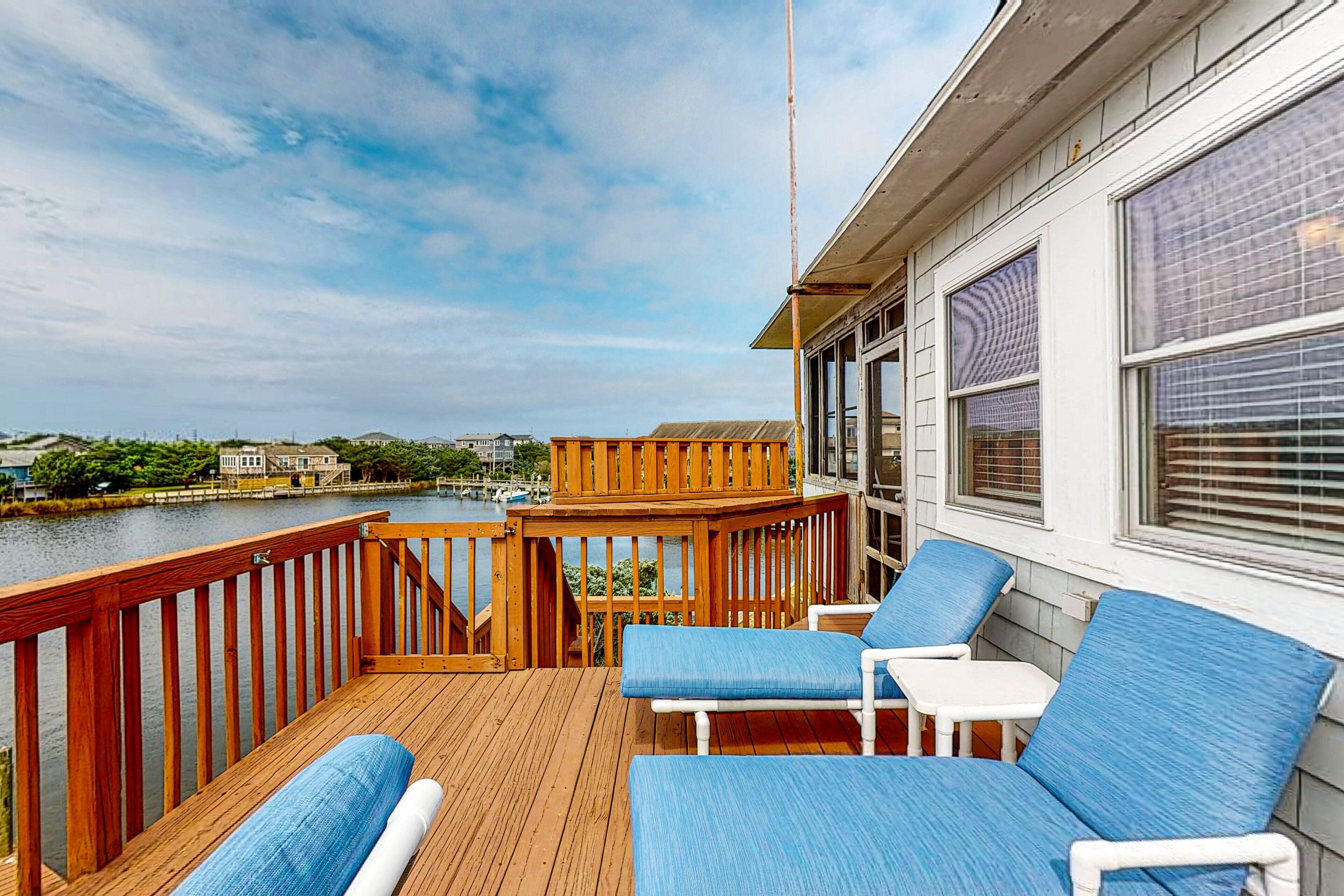 Une terrasse en bois avec des chaises rembourrées bleues et une petite table, donnant sur une étendue d’eau et un quai où sont amarrés des bateaux.