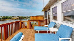 Une terrasse en bois avec des chaises rembourrées bleues et une petite table, donnant sur une étendue d’eau et un quai où sont amarrés des bateaux.