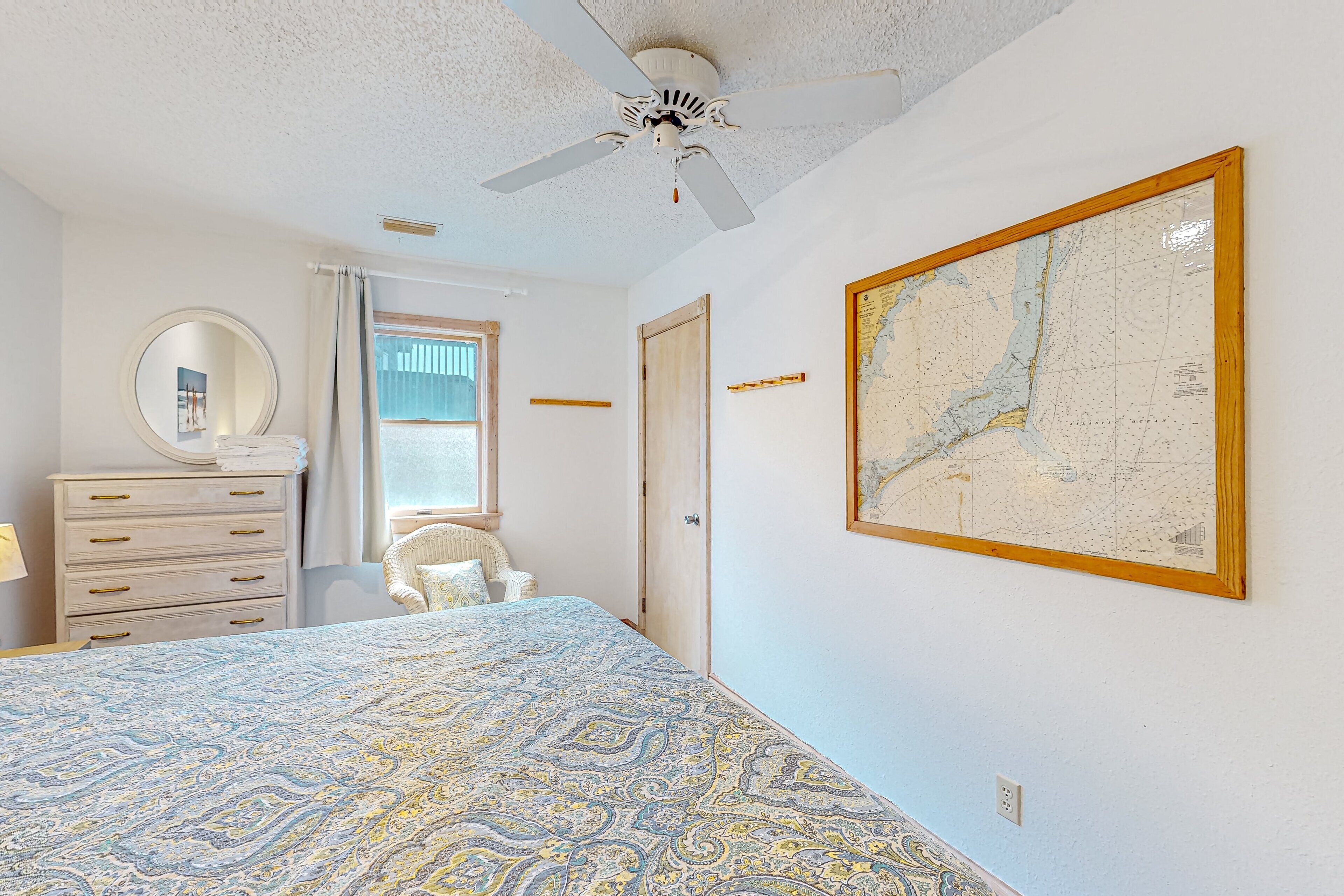 A bedroom with a patterned bedspread, a ceiling fan, a map on the wall, a dresser, and a chair.