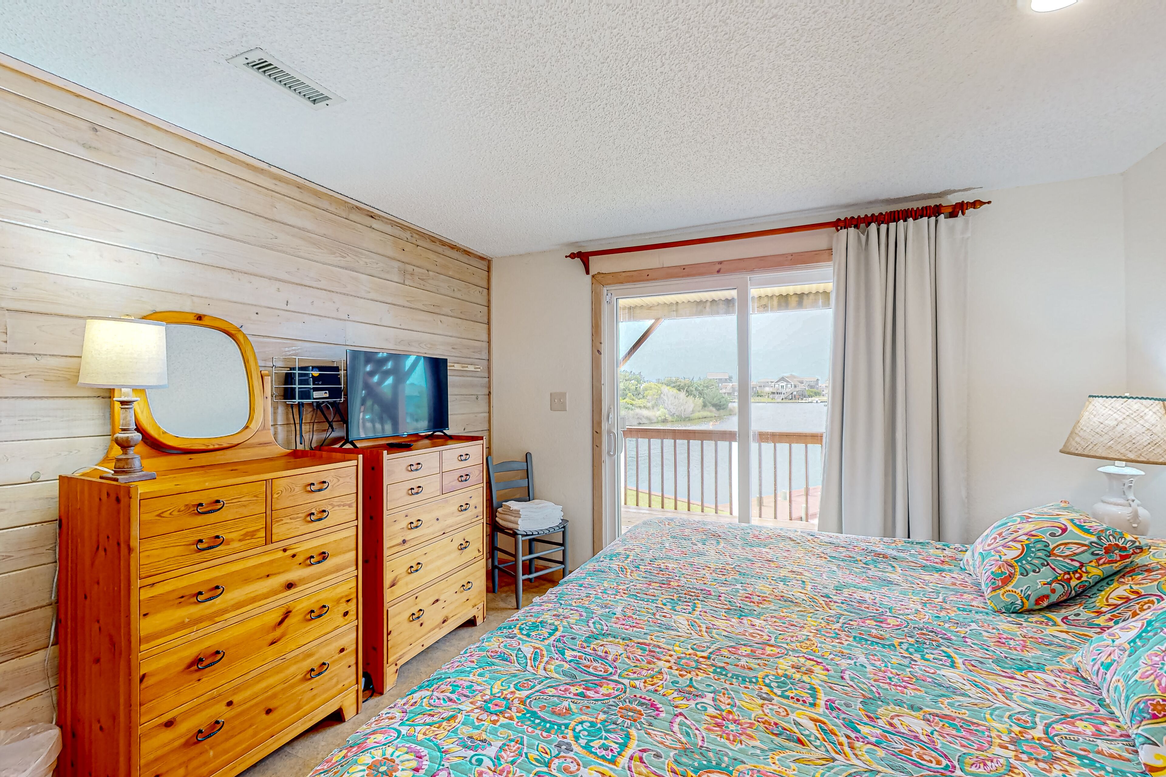A bedroom with a wooden dresser, a bed with a colorful patterned bedspread, a television on a wooden cabinet, and a view of a balcony with a railing.