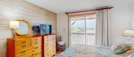 A bedroom with a wooden dresser, a bed with a colorful patterned bedspread, a television on a wooden cabinet, and a view of a balcony with a railing.