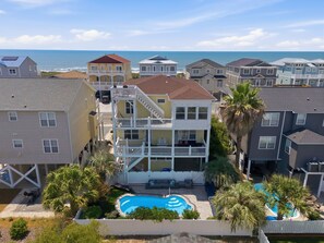 Um edifício residencial com vários andares, com piscina, rodeado por outras casas e uma praia ao fundo.