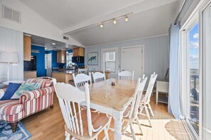 A spacious living area with a dining table surrounded by white chairs, a striped sofa, and a kitchen in the background.