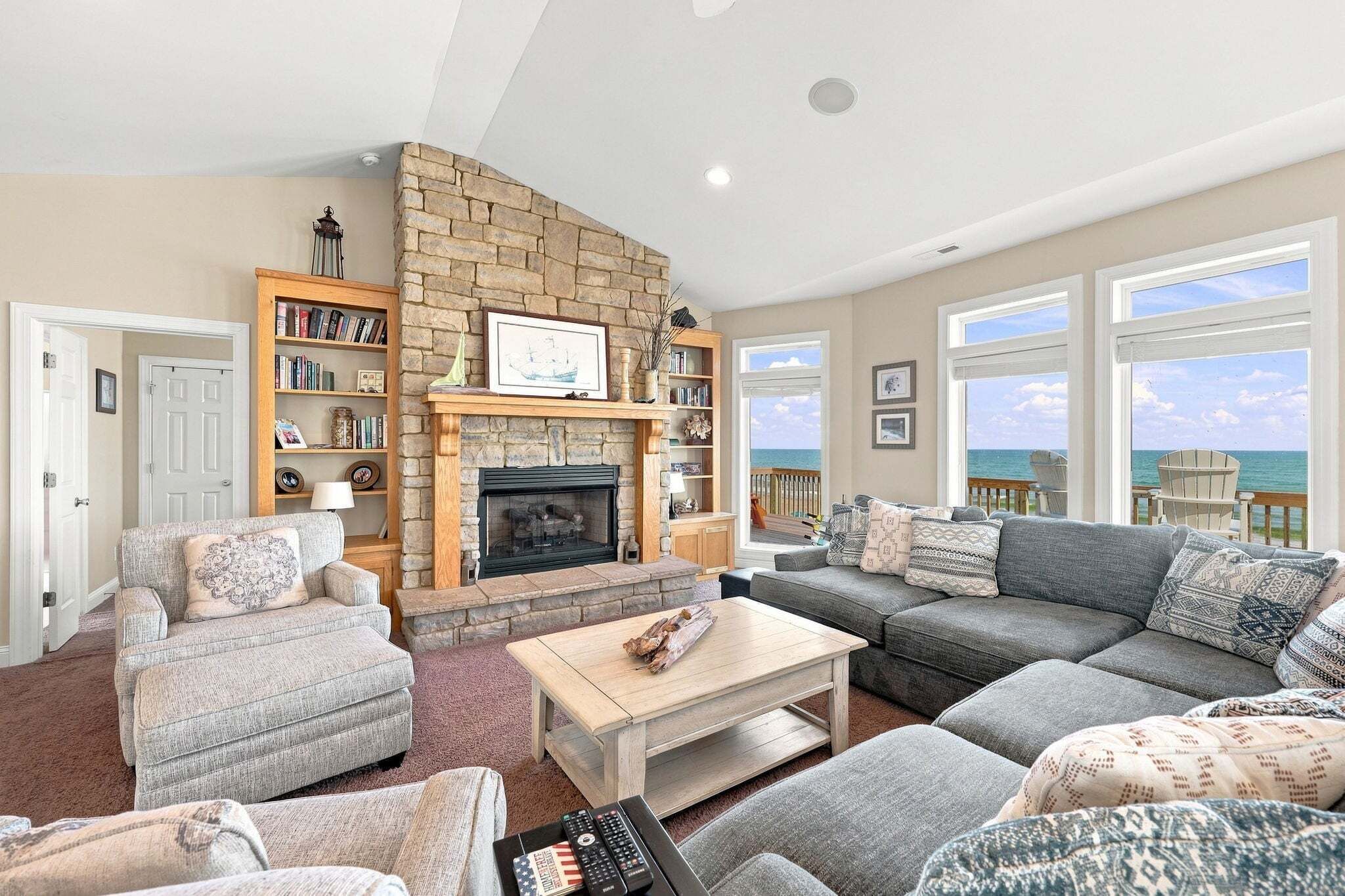 A living room with a stone fireplace, a bookshelf, a sofa, and a view of the ocean through large windows.