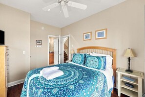 Une chambre à coucher comprenant un lit, une table de chevet, une lampe, un ventilateur de plafond et une porte donnant sur un escalier.