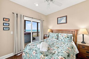Une chambre avec un lit, une table de chevet, une lampe, un ventilateur de plafond et une vue sur l’océan par une porte coulissante.