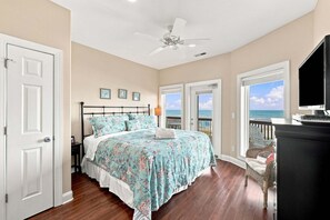 A bedroom with a bed, a ceiling fan, a TV, and a view of the ocean.