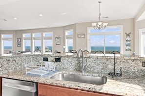 A kitchen with a granite countertop, stainless steel sink, and a view of the ocean through large windows.