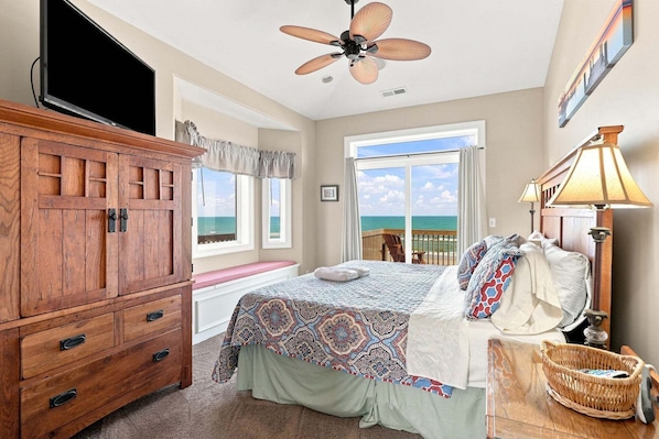 A bedroom with a bed, a wooden dresser, a ceiling fan, a window with a view of the ocean, and a lamp.