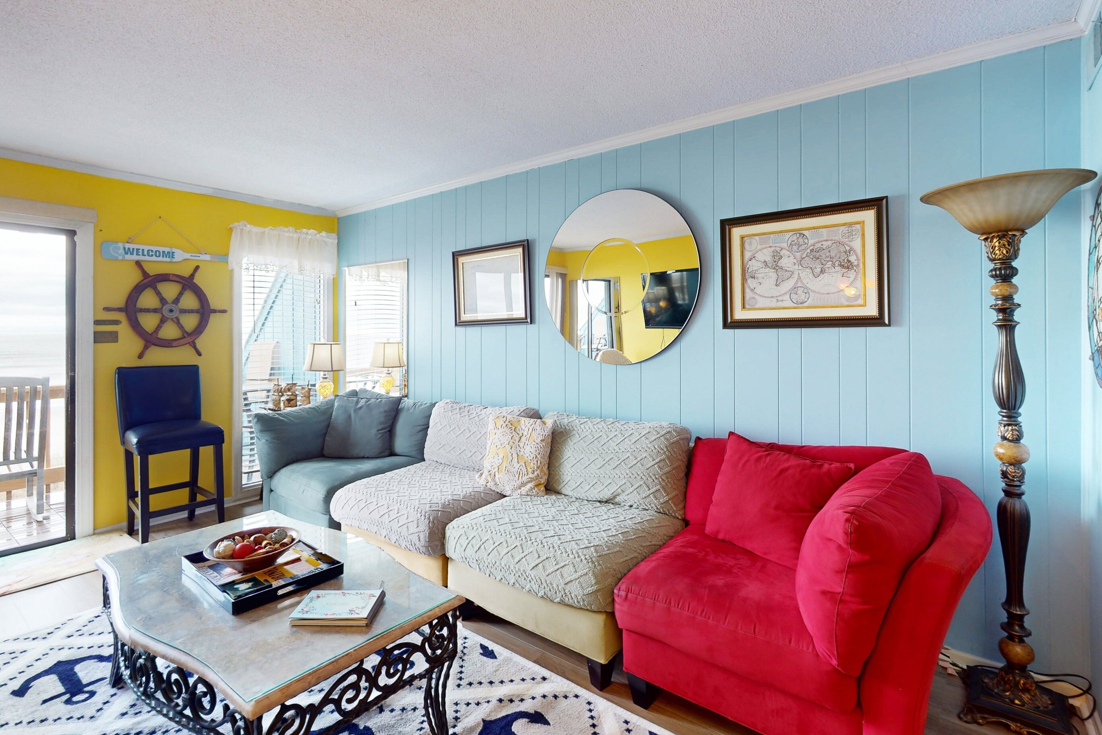 A living room with a blue wall, a red sofa, a beige couch, a glass coffee table, and a wooden chair.
