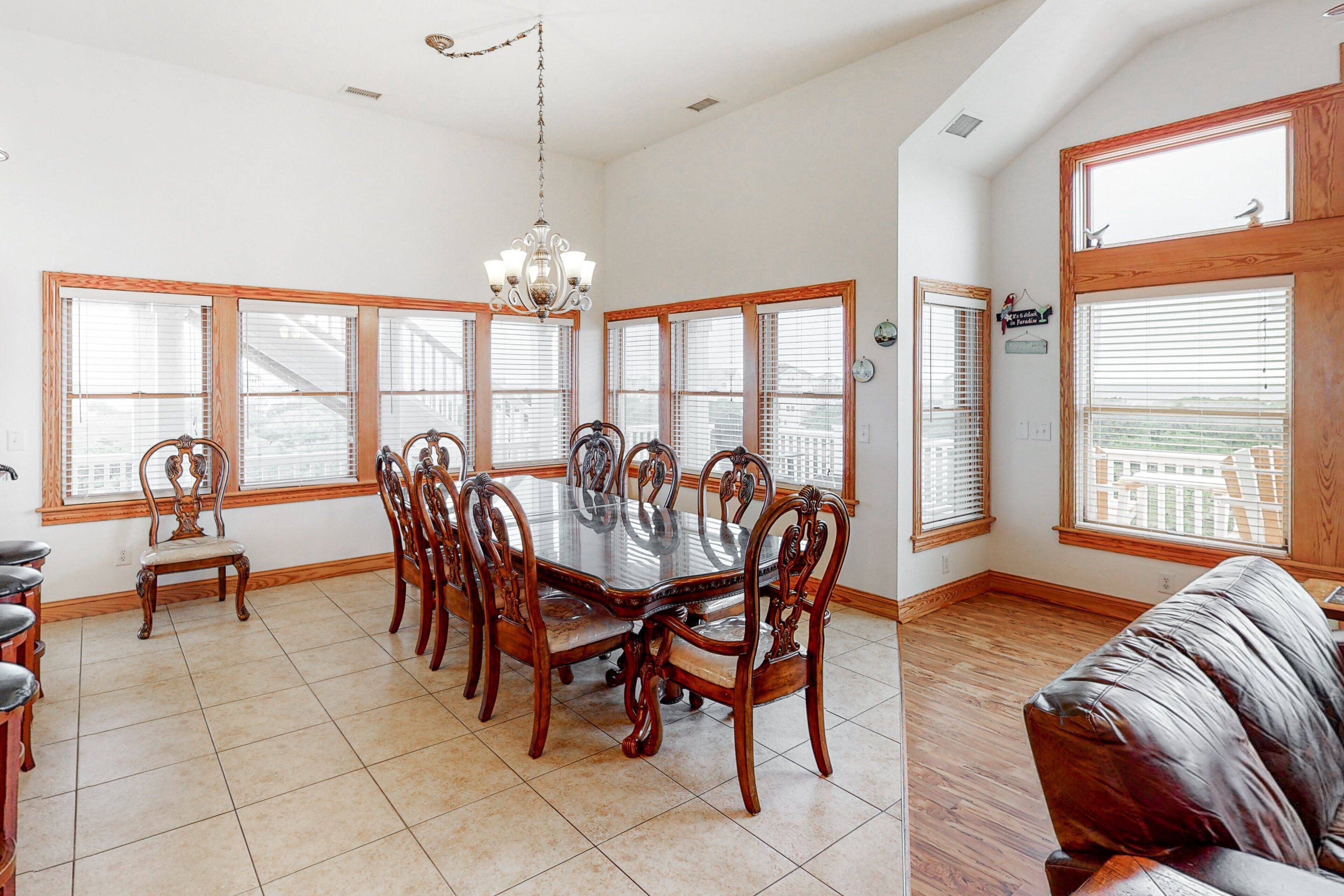 A dining area with a large wooden table surrounded by chairs, large windows with blinds, and a chandelier.