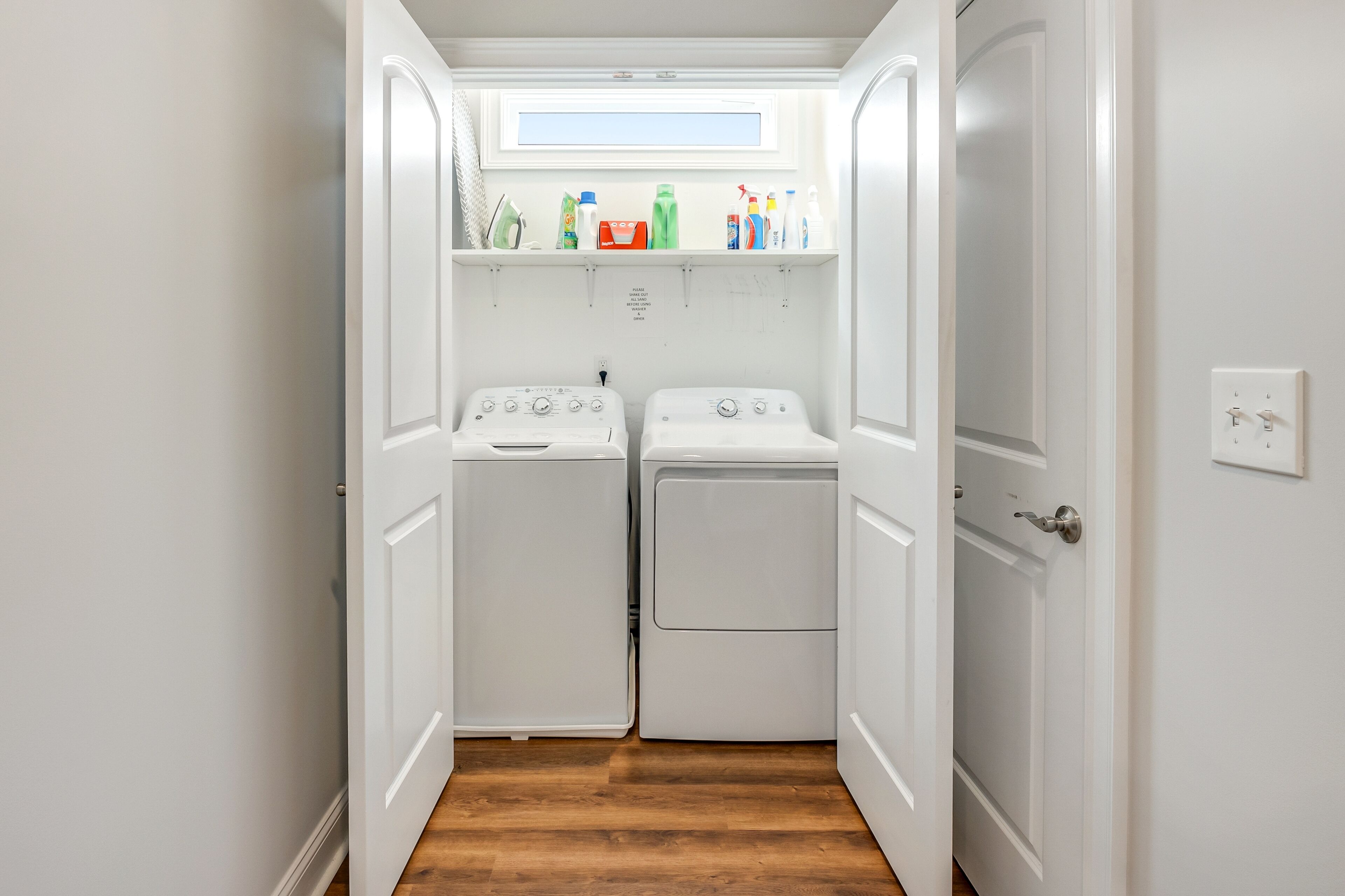 A laundry room with two washing machines and a shelf above containing cleaning supplies.