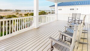 A wooden deck with white railings and a ceiling fan, overlooking a landscape with buildings and trees. - Take Me A-Wave: Water-view, pool, foosball, grill, & two W/Ds (Ocean Isle Beach)