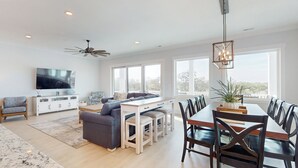 A spacious living room with a large TV, a dining area with a wooden table and chairs, and a ceiling fan. - Take Me A-Wave: Water-view, pool, foosball, grill, & two W/Ds (Ocean Isle Beach)
