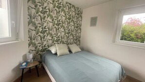 Une chambre à coucher avec un lit, une petite table sur laquelle se trouve une bouteille d’eau, un tableau encadré et une fenêtre donnant sur un paysage verdoyant.
