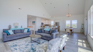 A spacious living area with a gray sofa, patterned armchairs, and a coffee table. A kitchen is visible in the background with modern appliances. - Ocean-view beach home - private hot tub, elevator, cook's kitchen - dog-friendly (North Topsail Beach)