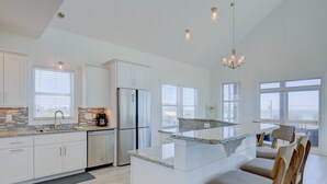 A modern kitchen with white cabinetry, a stainless steel refrigerator, and a granite countertop island with bar stools. - Ocean-view beach home - private hot tub, elevator, cook's kitchen - dog-friendly (North Topsail Beach)