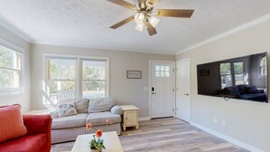Un soggiorno con un divano rosso, una poltrona beige e un tavolino da caffè con fiori. Si nota una ventola a soffitto, una TV montata a parete e una porta che dà su un'altra stanza.