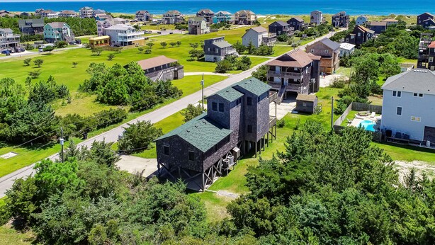 An aerial view of a coastal residential area with houses, a large building with a green roof, and a beach in the background. - Handsome raised home with private hot tub, & central AC (Waves)
