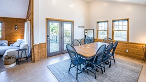 A spacious room with a large wooden dining table, multiple chairs, a sofa, and a small side table. There are two large windows with wooden frames and a sliding glass door leading to an outdoor deck. - Handsome raised home with private hot tub, & central AC (Waves)