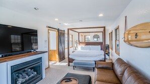 A modern bedroom with a large bed, a TV, a fireplace, and a surfboard on the wall. - Handsome raised home with private hot tub, & central AC (Waves)