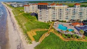 Un hotel frente a la playa con varios edificios altos, un área de piscina central y un acceso peatonal a la playa.