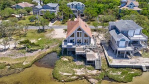 A large house with a prominent front porch and multiple decks, surrounded by a natural landscape with a small stream and sandy areas. - Waterfront  house with pool, W/D, private hot tub, & AC (Frisco)