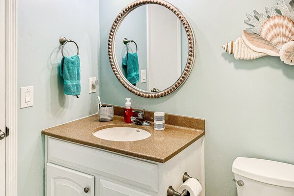 A bathroom with a sink, mirror, and toilet. There are towels hanging on the wall, a soap dispenser, and a toilet paper holder. - The Turquoise Pearl: Waterfront, pool, dock access (Ocean Isle Beach)