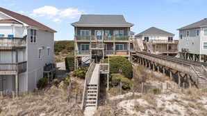 Une propriété située en bord de mer, avec un bâtiment principal doté d’un balcon et d’une passerelle en bois menant à celui-ci.