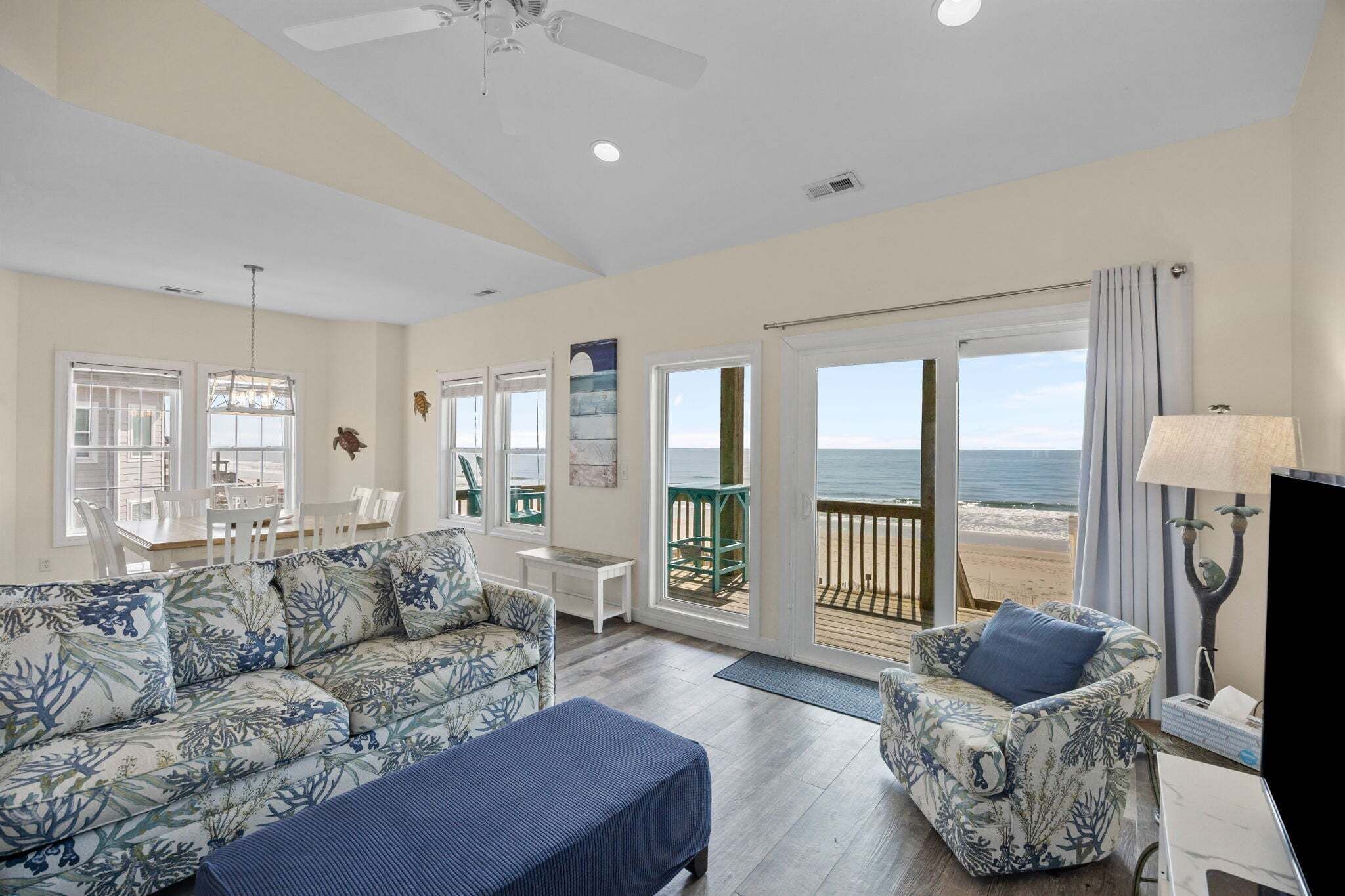 A living room with a sofa, armchair, and a TV stand. There is a balcony with a view of the beach.