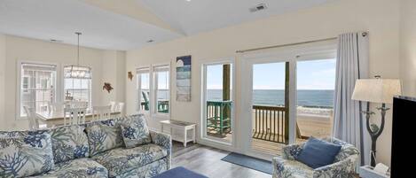A living room with a sofa, armchair, and a TV stand. There is a balcony with a view of the beach.