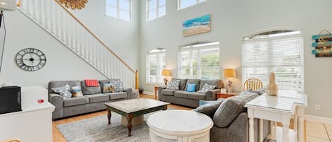 A spacious living room with a staircase, large windows, and a ceiling fan.