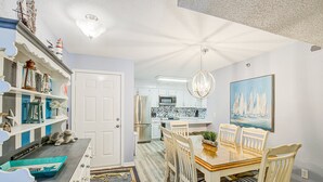 Un coin repas avec une table et des chaises en bois, une cuisine en arrière-plan et une étagère décorative où sont exposés divers objets.