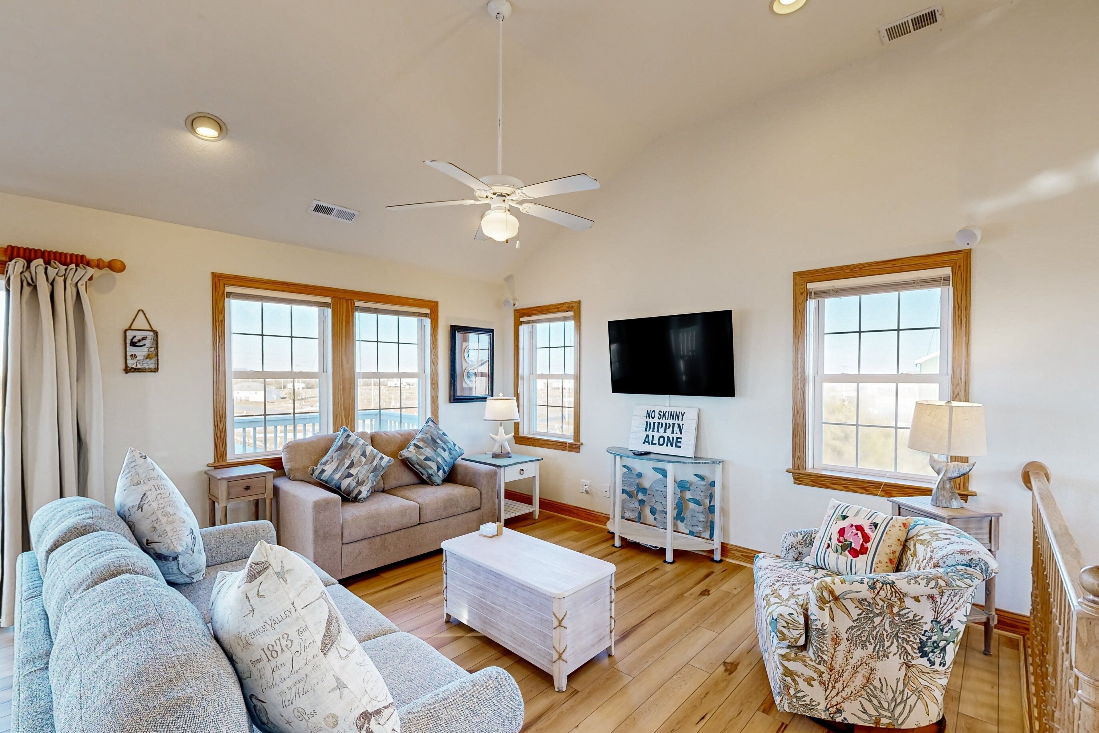 A spacious living room with a ceiling fan, large windows, a flat-screen TV, and a cozy seating area with patterned cushions.
