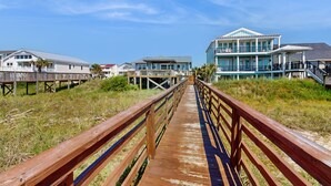 A wooden boardwalk leading to a beachfront property with multiple buildings, including a large house with a balcony. - Deja Blue: Oceanfront Perfection (Ocean Isle Beach)