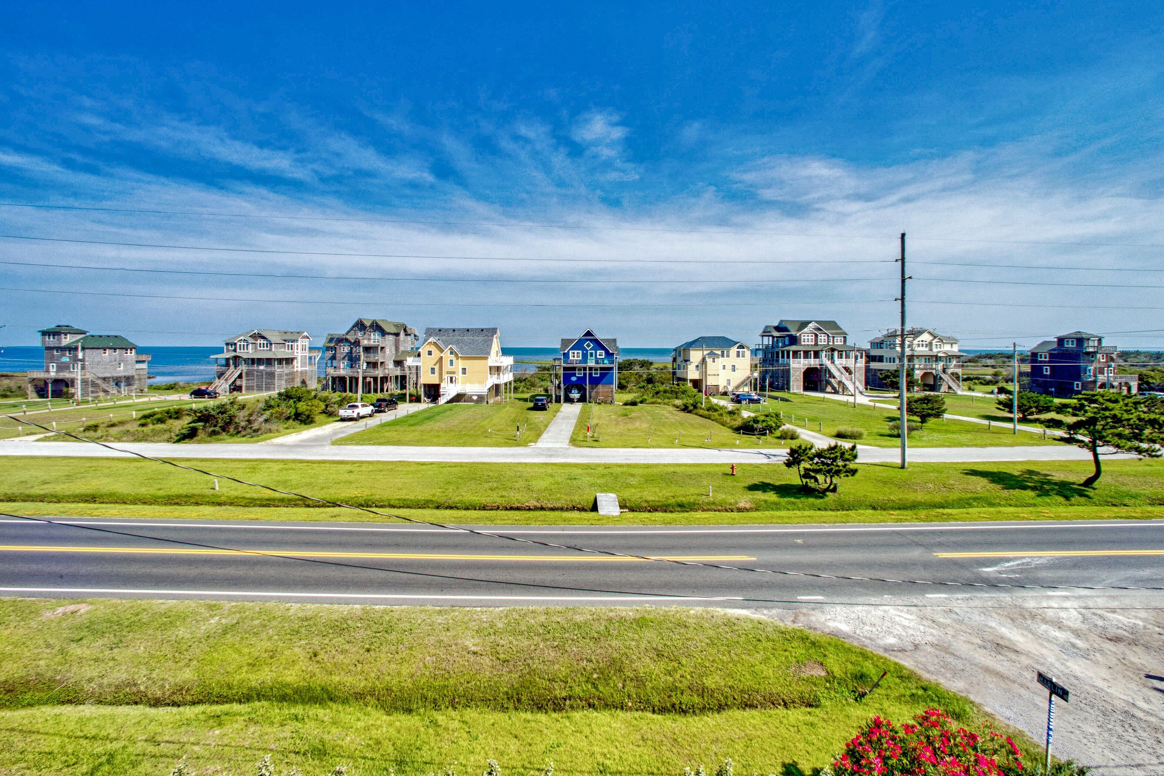 A coastal residential area with houses featuring multiple stories and balconies, a clear road, and a view of the ocean.
