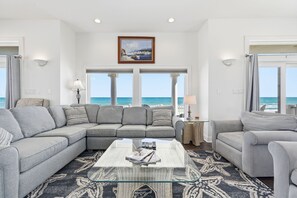 A living room with a large sectional sofa, a glass coffee table, and a view of the ocean through large windows.