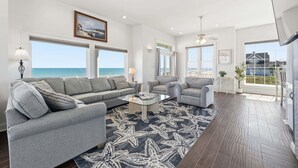 A spacious living room with a large window overlooking the ocean, a gray sofa, a coffee table, and a ceiling fan. - Oceanfront, Next door to Pool, Easy Access, Oversized Deck (North Topsail Beach)
