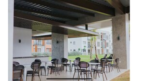 Un espace extérieur aménagé, abrité et doté de chaises et de tables, un design architectural moderne avec des piliers en béton, et une vue sur des immeubles résidentiels en arrière-plan.