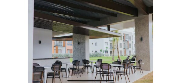 A covered outdoor seating area with chairs and tables, a modern architectural design with concrete pillars, and a view of residential buildings in the background. - Charming 2-bedroom condo with fitness room and AC in Punta Cana (Punta Cana)