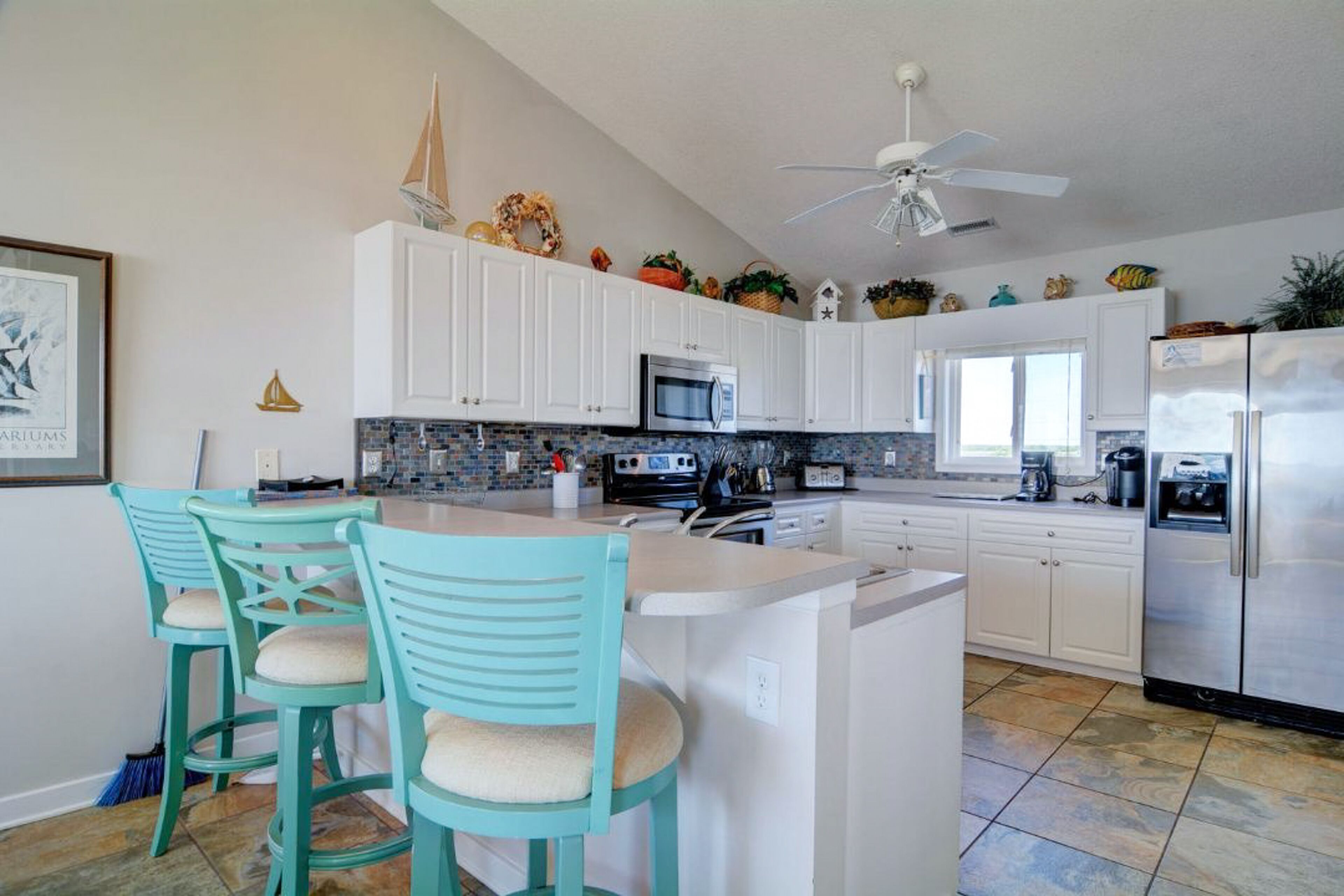 A modern kitchen with white cabinets, a central island with bar stools, a ceiling fan, and decorative items on the upper cabinets.