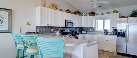 A modern kitchen with white cabinets, a central island with bar stools, a ceiling fan, and decorative items on the upper cabinets.