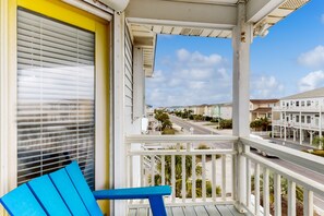 A balcony with a blue chair, a white railing, and a view of a street with buildings. - Sea-Rendipity: Dog-friendly, across beach, pool (Ocean Isle Beach)